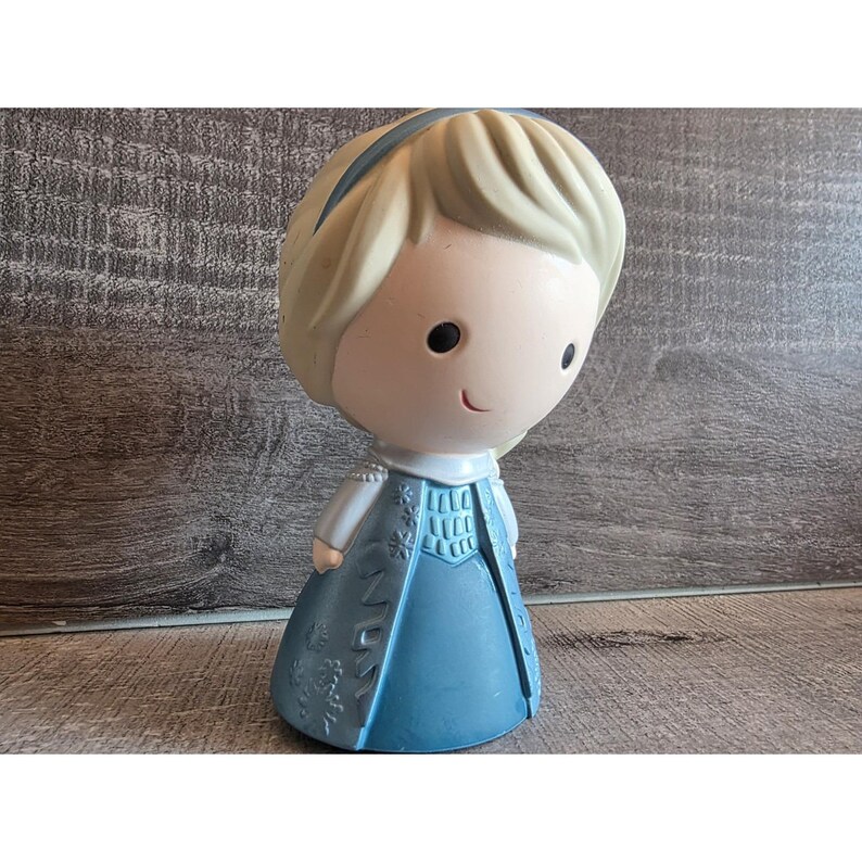 May include: A figurine of a cartoon character with a light blue dress and a blonde hairstyle. The figurine has a small smile and a blue headband. The dress has a detailed design. The figurine is standing on a wooden surface.