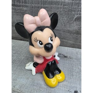 May include: A Minnie Mouse figurine with a pink bow, black ears, and a red dress. The figurine has yellow shoes and white gloves. The figure is sitting down and has a cheerful expression.