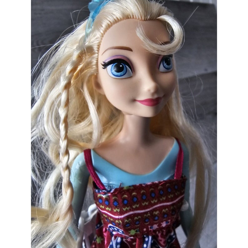Puede incluir: Una mu&ntilde;eca rubia con ojos azules y un vestido estampado en rojo y blanco. La mu&ntilde;eca lleva una diadema azul en el pelo.
