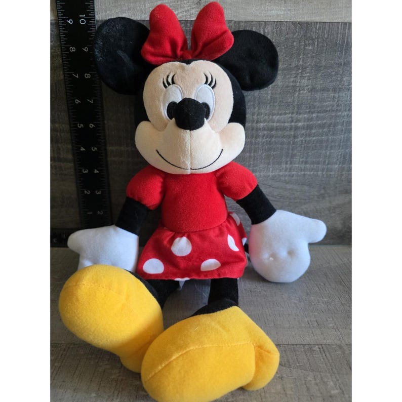 May include: A plush Minnie Mouse doll. The doll has a red dress with white polka dots, a red bow, black ears, and yellow shoes. The doll's arms and hands are white, and the face is beige with a black nose and smiling mouth.