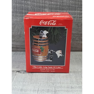 May include: A red Coca-Cola box featuring a decorative ornament. The ornament depicts a wooden barrel with a Coca-Cola logo, a tap, and two white mice. The text on the box reads "The Cold, Crisp Taste Of Coke".