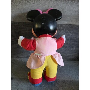 May include: A vintage Minnie Mouse plush doll, viewed from the back. The doll has a black head with large ears, a pink bow, and is wearing a red and pink outfit with yellow pants. The doll's shoes are red with blue soles.