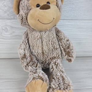 May include: A plush monkey toy with shaggy light brown fur, a smooth tan face, dark brown embroidered eyes, and a stitched smile. The stuffed animal features rounded ears, long arms, and tan-colored feet, crafted from soft, textured fabric.
