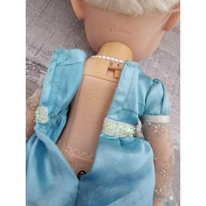 May include: A light blue dress with a glittery silver belt and a white lace sleeve. The doll has blonde hair and is wearing a pearl necklace. The back of the doll shows a battery compartment with an on/off switch and the text "&copy;DISNEY".