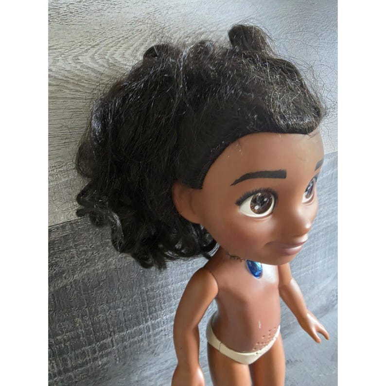 May include: A doll with dark skin, brown eyes, and black hair styled in a ponytail. The doll is wearing a white diaper and a blue necklace. The doll's arms are outstretched, and it is looking to the side.