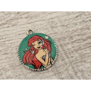 May include: A round, silver charm with a green background and a colorful image of Ariel, the Little Mermaid, with red hair and a seashell in her hand.