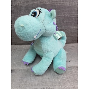 Puede incluir: Un peluche de dragón turquesa con detalles morados en los pies, espinas y orejas. El dragón tiene grandes ojos azules, un diente blanco y pequeñas alas verdes. El juguete está hecho de un material suave y difuso.