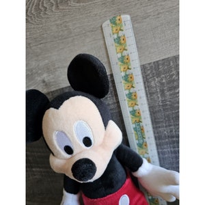 Puede incluir: Un juguete de peluche Mickey Mouse negro y rojo con grandes ojos blancos y una nariz negra. El juguete mide aproximadamente 50 cm de alto y est&aacute; sentado sobre una superficie de madera.