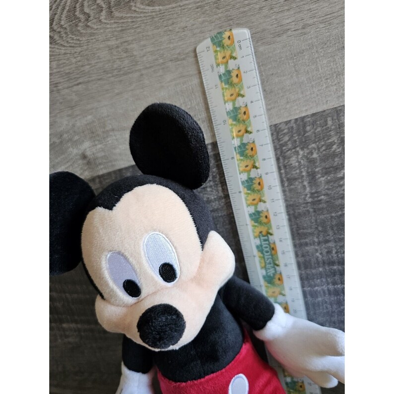 Puede incluir: Un juguete de peluche Mickey Mouse negro y rojo con grandes ojos blancos y una nariz negra. El juguete mide aproximadamente 50 cm de alto y est&aacute; sentado sobre una superficie de madera.