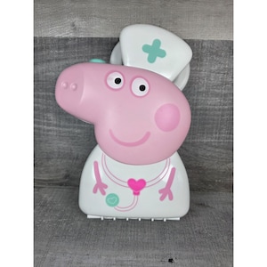 Puede incluir: Un juguete rosa de Peppa Pig con un disfraz de enfermera. El juguete tiene un sombrero blanco con una cruz turquesa, una bata blanca con un corazón rosa y un diseño de estetoscopio, y una cara sonriente. El juguete es de plástico.