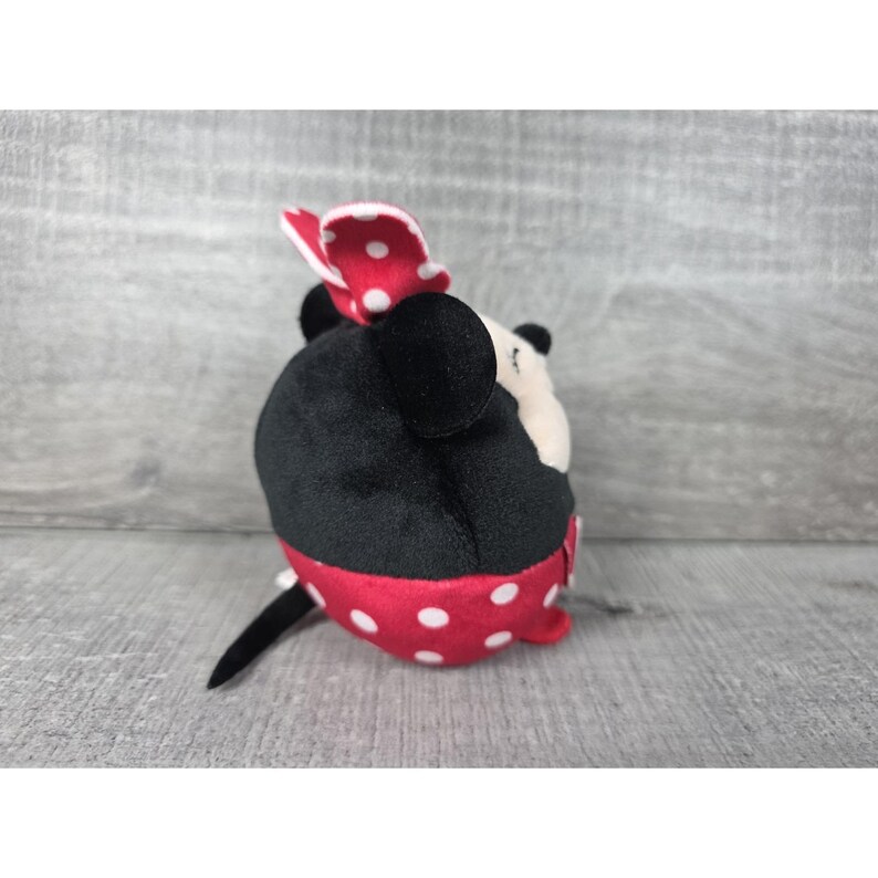 May include: A plush toy shaped like a mouse with a round black body, a red skirt featuring white polka dots, and a matching red and white polka dot bow on top. The toy has black ears, a small black tail, and a cream-coloured face with visible stitching.