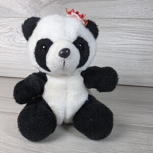 May include: A plush panda bear toy with black and white fur, featuring black patches around the eyes, black ears, arms, and legs, and a white face and torso. A small, frayed red and white fabric accent is attached to the top of the panda's head.