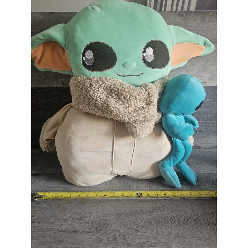 May include: A large green plush toy with a brown fur scarf and big eyes. The toy is sitting on a wooden surface with a smaller blue plush toy sitting on its back.