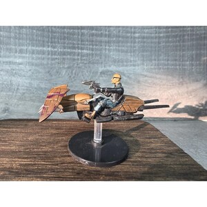 May include: A miniature figurine of a futuristic vehicle with a rider. The vehicle is primarily brown and purple, with sleek lines and a wing-like structure. The rider is wearing a black outfit and holding two guns. The figurine is mounted on a black base.
