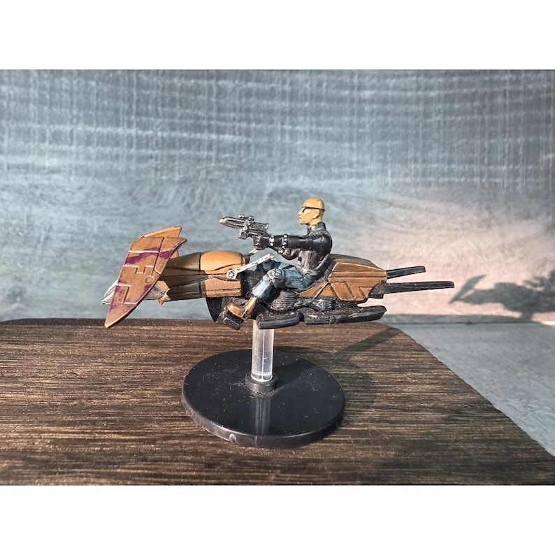 May include: A miniature figurine of a futuristic vehicle with a rider. The vehicle is primarily brown and purple, with sleek lines and a wing-like structure. The rider is wearing a black outfit and holding two guns. The figurine is mounted on a black base.