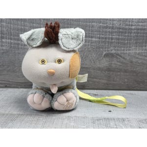 May include: A plush toy dog with gray fur, floppy ears, and a brown tuft of hair. The dog has tan patches on its face and paws, with yellow eyes and a pink nose. A yellow ribbon is attached.