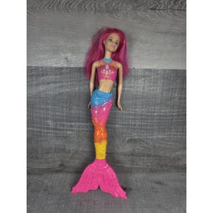 Może przedstawiać: Lalka syrenka z różowymi włosami i kolorowym ogonem. Góra lalki jest różowa z dekoracyjnym wzorem, a ogon przechodzi od niebieskiego do różowego, pomarańczowego i żółtego. Lalka stoi na szarej drewnianej powierzchni.
