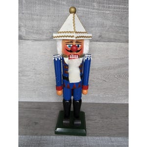 May include: A wooden nutcracker with a blue and red uniform, a white hat with gold trim, and a white beard. The nutcracker is standing on a green base.