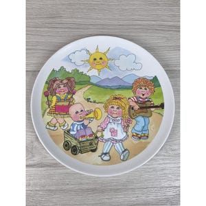 Może przedstawiać: Okrągły biały talerz ceramiczny z kolorową ilustracją czworga dzieci grających na instrumentach na zewnątrz. Scena przedstawia słońce na niebie, zielone wzgórza i polną drogę. Dzieci są przedstawione z różnymi instrumentami, w tym bębnem, rogiem i gitarą, a jedno z nich siedzi w małym drewnianym wózku.