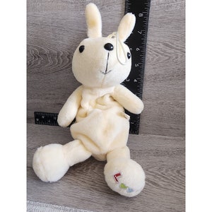 Puede incluir: Un juguete de peluche de conejo blanco con ojos negros y un pequeño lazo en la cabeza. El conejo está sentado con las piernas extendidas y tiene un pequeño diseño bordado en un pie.