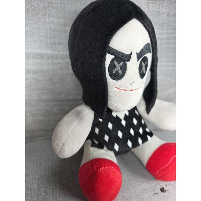 May include: A plush doll with a white face, black hair, and a black and white diamond-patterned dress. The doll has button eyes with an X design and red shoes. The doll's facial expression is stern.