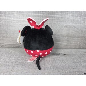 May include: A round plush toy featuring a black upper section and a red lower section with white polka dots. The toy has two black ears, a red and white polka-dot bow on top, and a thin black tail extending from the bottom. A white tag is attached.