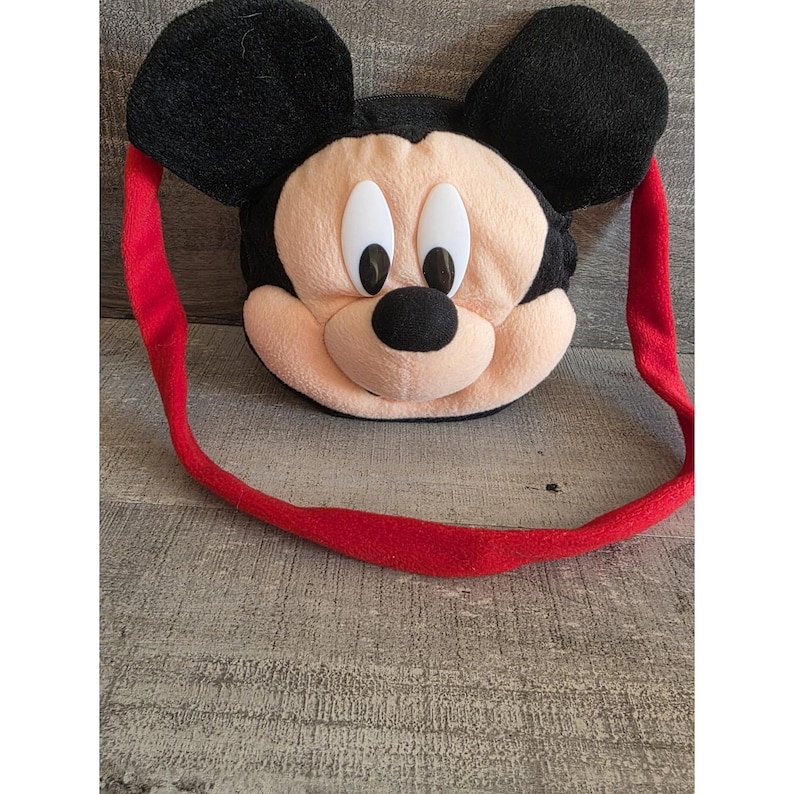 May include: A plush Mickey Mouse-shaped bag with a red strap. The bag features a black head and ears, a tan face, and a black nose. The strap is a bright red color.