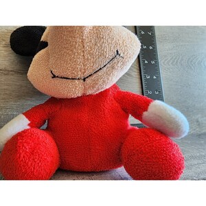 以下が含まれることがあります： 黒い鼻と白い腕と脚を持つ赤いぬいぐるみ。おもちゃは縫い付けられた笑顔をしていて、木製の表面に座っています。