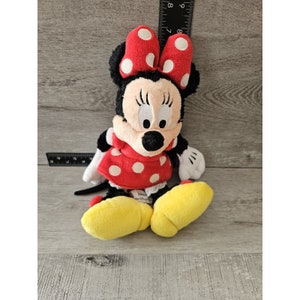 K&ouml;nnte beinhalten: Ein rotes und wei&szlig; gepunktetes Pl&uuml;schtier von Minnie Maus. Sie tr&auml;gt ein rotes Kleid mit wei&szlig;en Punkten und eine rote Schleife mit wei&szlig;en Punkten. Sie hat gelbe Schuhe.