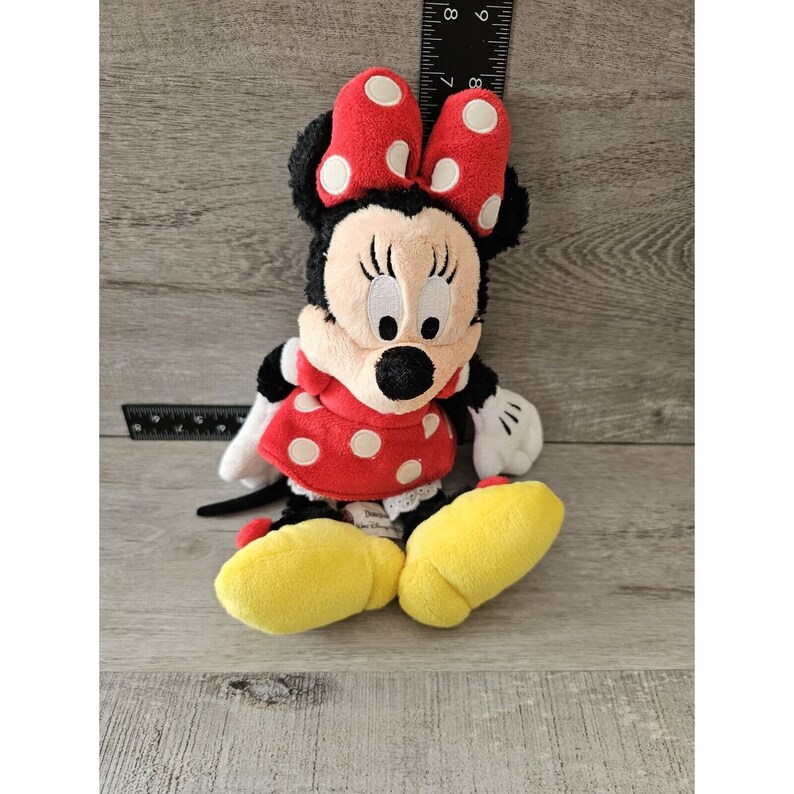 K&ouml;nnte beinhalten: Ein rotes und wei&szlig; gepunktetes Pl&uuml;schtier von Minnie Maus. Sie tr&auml;gt ein rotes Kleid mit wei&szlig;en Punkten und eine rote Schleife mit wei&szlig;en Punkten. Sie hat gelbe Schuhe.