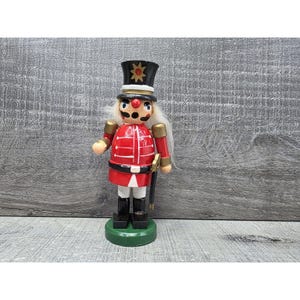 Puede incluir: Figura de soldado cascanueces de madera. La figura viste una chaqueta roja con detalles blancos, botas negras y un sombrero negro con una estrella dorada. Tiene barba blanca y está sobre una base verde.