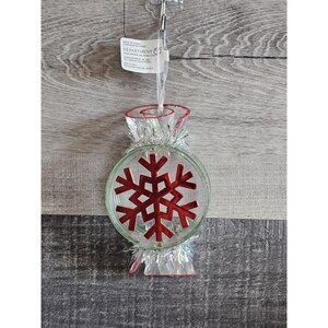 May include: A clear glass Christmas ornament shaped like a candy cane with a red snowflake inside. The ornament is decorated with silver tinsel.