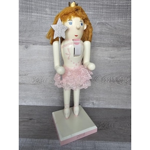 Puede incluir: Un cascanueces de madera blanco en forma de bailarina con un tutú rosa y una varita de estrella plateada. El cascanueces lleva una corona dorada y una cinta rosa alrededor del cuello. La base del cascanueces es blanca con un borde rosa y el año 2010 impreso en ella.