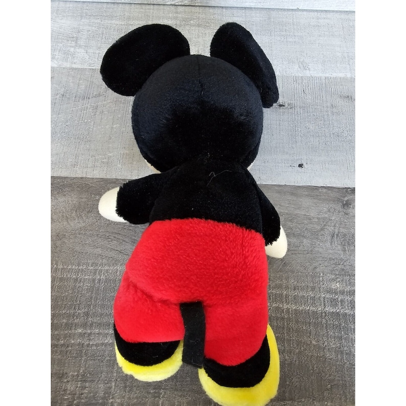 May include: A black and red plush Mickey Mouse toy. The toy is lying on its back with its arms and legs outstretched.