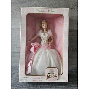 Puede incluir: Una muñeca Barbie con un vestido blanco con detalles rosas y un diseño floral. La muñeca está en una caja rosa y blanca con las palabras "Birthday Wishes" y "Collector Edition First in a Series" en ella.