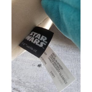 May include: A white and teal plush toy with a black tag that reads "STAR WARS" and "& TM Lucasfilm Ltd.". The tag also includes information about the toy's care, including "Surface Washable" and "Made in China".