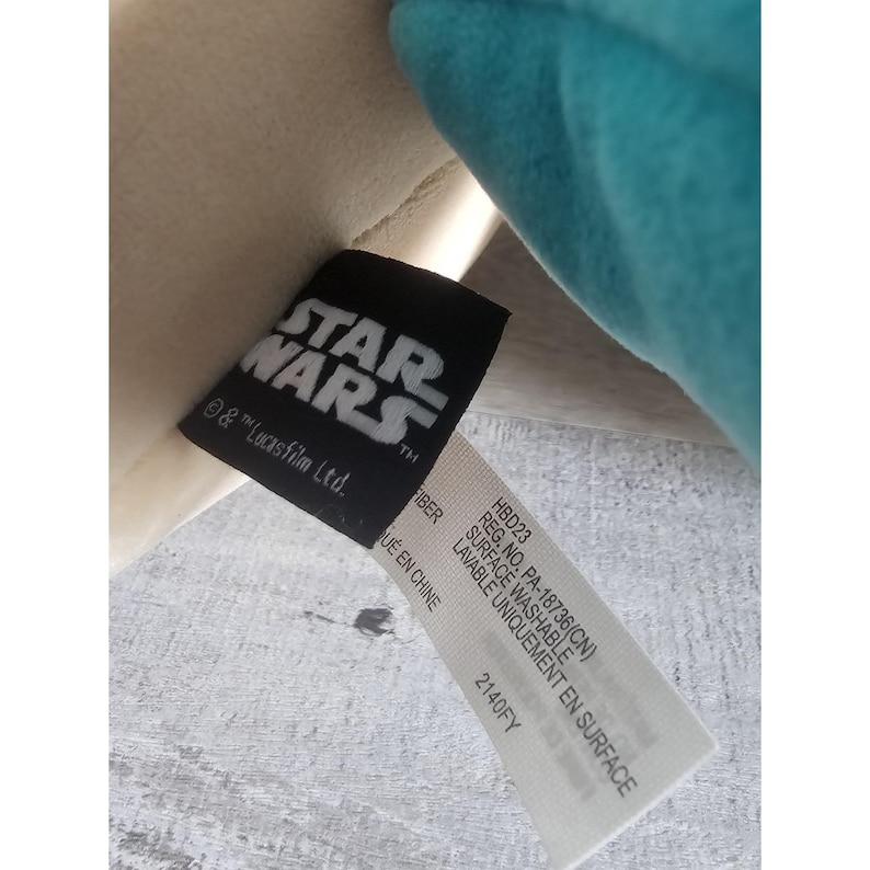 May include: A white and teal plush toy with a black tag that reads "STAR WARS" and "& TM Lucasfilm Ltd.". The tag also includes information about the toy's care, including "Surface Washable" and "Made in China".
