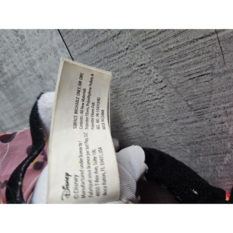 May include: A white tag with black text that reads "Disney" and "Surface Washable Only. Air Dry." The tag also includes information about the contents of the toy, including "Polyester Fibers, Polyethylene Pellets & Polyester Fibers Felt." The tag also includes the registration number "PA-15431 (HK)" and states that the toy was "Made in China."