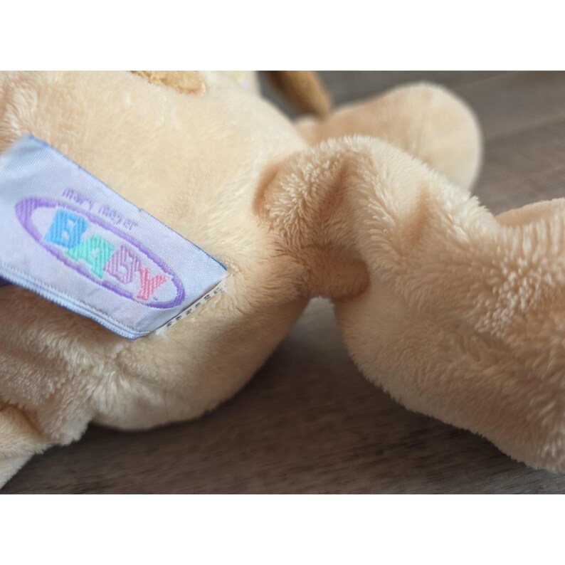 Puede incluir: Un peluche de color beige claro con una textura suave y difusa. Una etiqueta azul con la palabra "Baby" en letras coloridas est&aacute; adjunta. El juguete parece nuevo y sin usar.