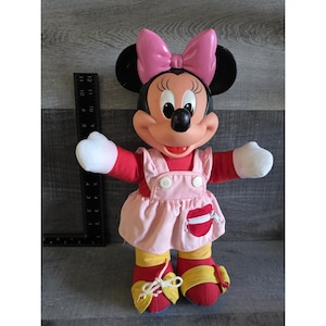 May include: A Minnie Mouse plush doll with a pink bow, black ears, and a red and pink outfit. The doll has white gloves, yellow and red shoes, and a smiling expression. The doll is approximately 12 inches tall.