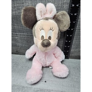 May include: A plush Minnie Mouse doll in a seated position. The doll has a pink body, a light pink face, and brown ears. The doll is approximately 8 inches tall.