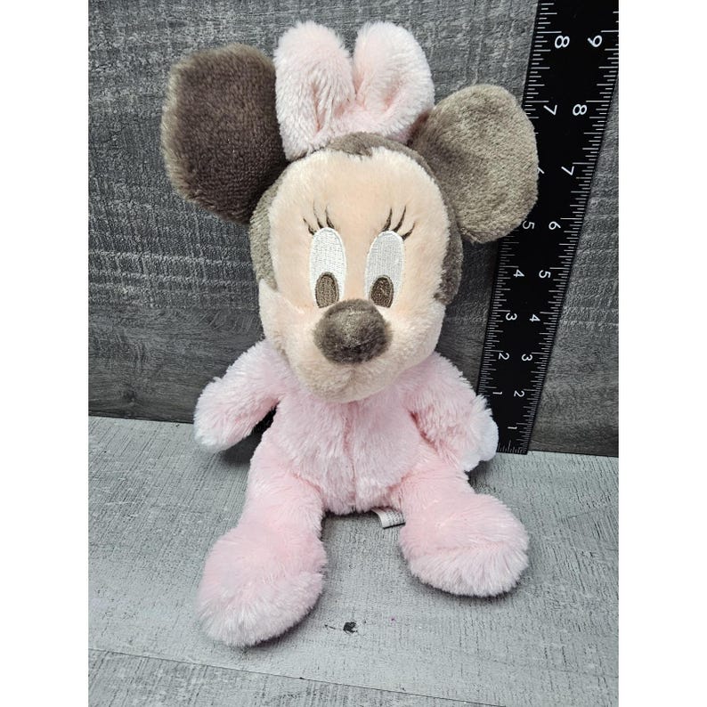 May include: A plush Minnie Mouse doll in a seated position. The doll has a pink body, a light pink face, and brown ears. The doll is approximately 8 inches tall.