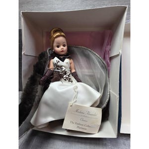 May include: A Madame Alexander Classics Platinum doll in its box. The doll is dressed in a white satin gown with a black and brown embroidered bodice, brown gloves, and a fur stole. The tag reads "Madame Alexander Classics The Fashion Collection Platinum."