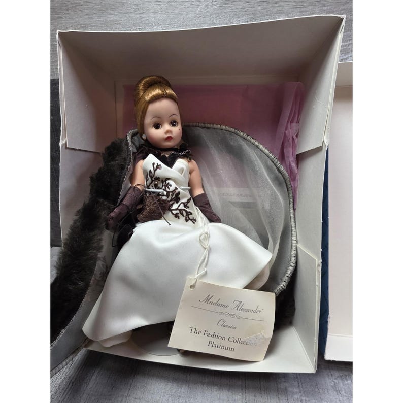 May include: A Madame Alexander Classics Platinum doll in its box. The doll is dressed in a white satin gown with a black and brown embroidered bodice, brown gloves, and a fur stole. The tag reads "Madame Alexander Classics The Fashion Collection Platinum."