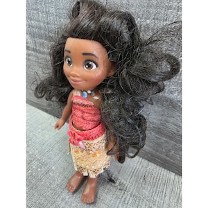 Op de afbeelding: Pop van een jong Polynesisch meisje met een donkere huid, bruine ogen en lang, zwart golvend haar. Ze draagt een rode en beige outfit met een ketting. De pop staat op een grijs oppervlak.