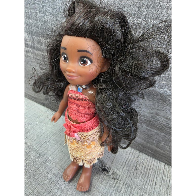 Op de afbeelding: Pop van een jong Polynesisch meisje met een donkere huid, bruine ogen en lang, zwart golvend haar. Ze draagt een rode en beige outfit met een ketting. De pop staat op een grijs oppervlak.