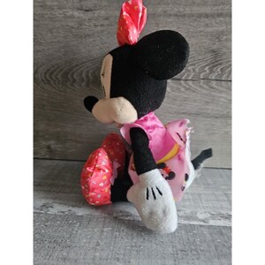 May include: A plush Minnie Mouse toy wearing a pink dress with white polka dots and a red bow. The toy is sitting with its legs crossed and its arms outstretched.