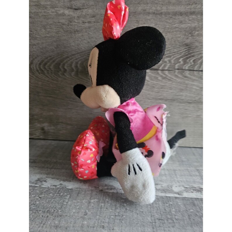 May include: A plush Minnie Mouse toy wearing a pink dress with white polka dots and a red bow. The toy is sitting with its legs crossed and its arms outstretched.