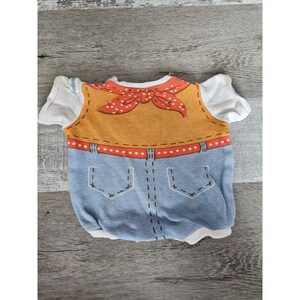 May include: A blue and yellow baby onesie with a cowboy design. The onesie has a red bandana, a brown vest, a blue denim bottom, and a red belt with white polka dots.