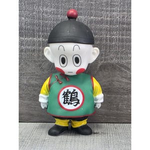 May include: A figurine of a character with a black cap and a red sphere on top. The character has large eyes, red cheeks, and a green vest with a circular emblem. The figure is wearing yellow sleeves and pants, with black shoes.